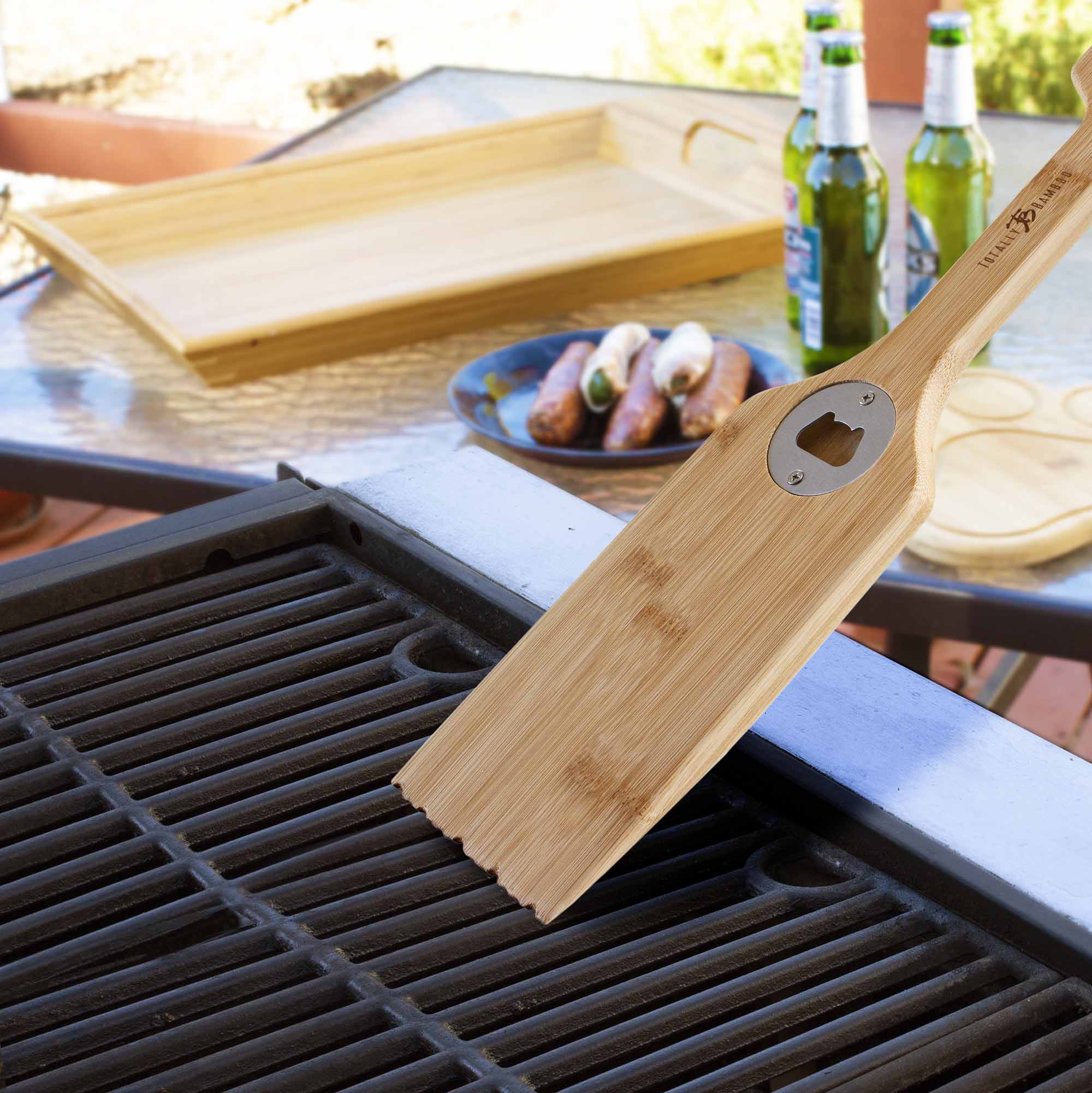 A Totally Bamboo BBQ Grill Scraper with a bottle opener rests on a barbecue grill. In the background, a tray, a plate of sausages, and two beer bottles sit on an outdoor patio table.