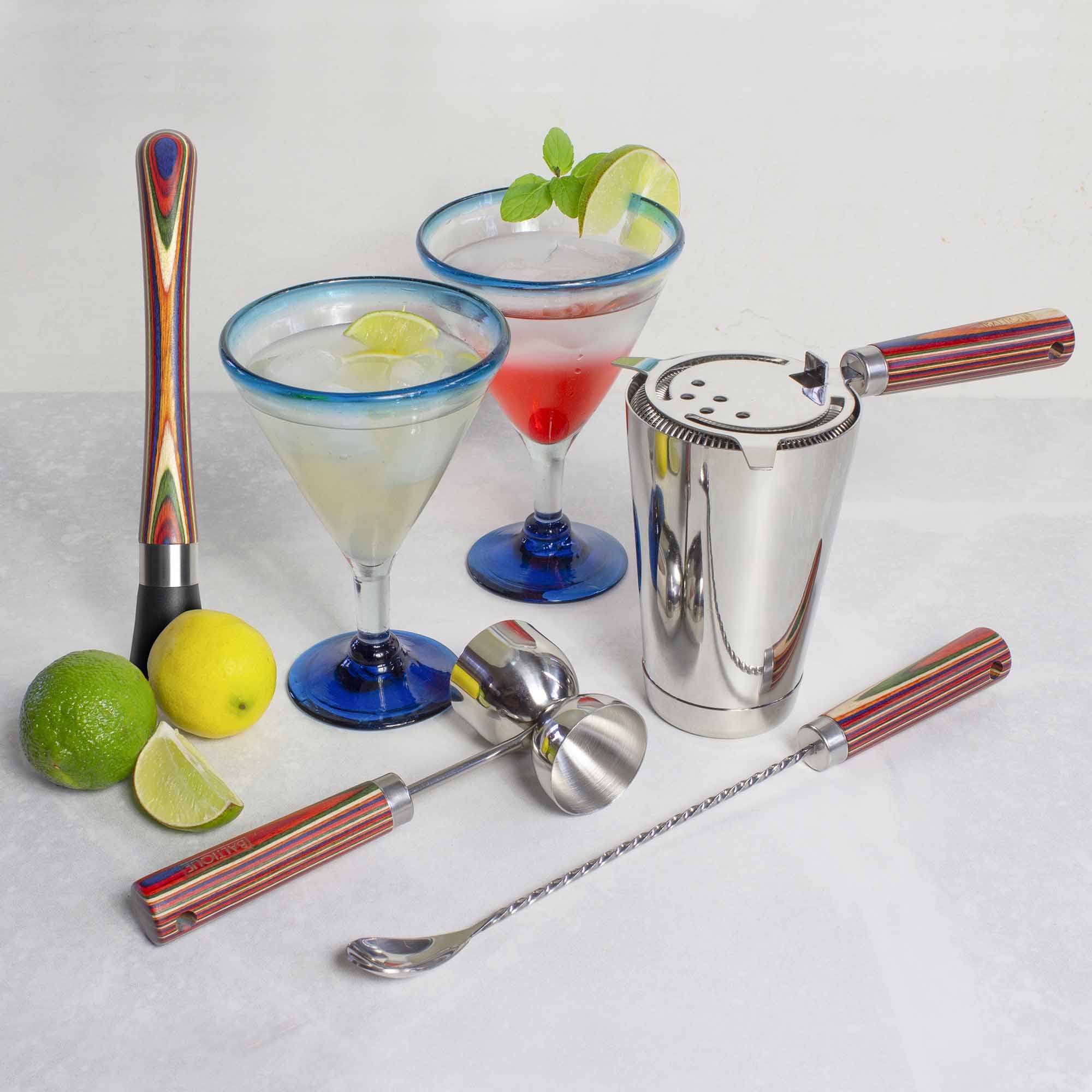The Totally Bamboo Baltique® Marrakesh Collection 4-Piece Bartending Kit includes a colorful muddler, jigger, shaker, and spoon. On a marble surface are two martini glasses, one lime and one red cocktail with lime and mint garnishes nearby lemons and limes.
